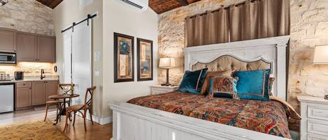 Un dormitorio con una cama grande, una pared de piedra, techo de madera y una cocina al fondo.