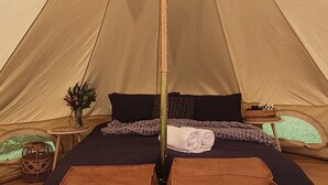 Teltta, jossa on keskellä tukipylväs, sänky tummalla peitolla, kaksi ruskeaa nahkaista jalkarahi ja pieni pöytä, jossa on maljakko ja kori.