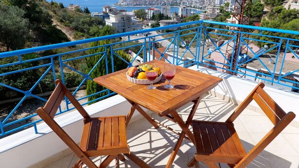 A balcony with a wooden table and chairs, a bowl of fruit, and two glasses of red wine. The balcony overlooks a cityscape with buildings and a view of the sea. - Charming 2-Bed Apartment in Sarandë (Sarande)