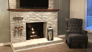 A living room with a brick fireplace, a flat-screen TV mounted above it, and a dark-colored armchair.