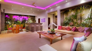 A modern living room with a sofa, coffee table, and chairs. There is a bar area with stools and a plant. The room has a ceiling fan and is decorated with plants and flowers. - Elegant Private Villa, 3 BR, Seminyak With Staff (Keroboken Kelod)