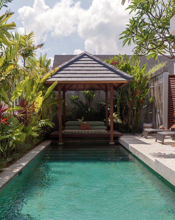 A pool with a wooden gazebo and a bench, surrounded by tropical plants.