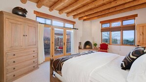 A bedroom with a wooden ceiling, a fireplace, a large bed, and a view through the glass doors.