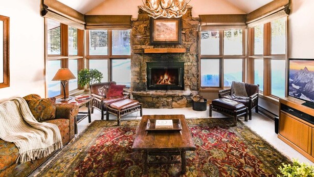 A cozy living room with a stone fireplace, comfortable seating, and large windows offering a view of the outdoors.