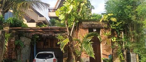 Una casa tradicional con un coche estacionado frente a ella, rodeada de abundante vegetación y un tejado de paja.