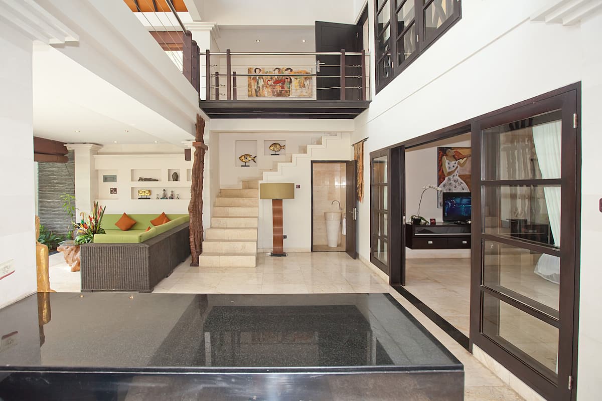Un intérieur spacieux avec un escalier menant à un niveau supérieur, un coin salon avec des coussins, une grande fenêtre et un téléviseur à écran plat.