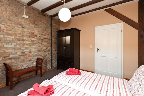 A room with a brick wall, a wooden bench, a dark wooden wardrobe, a white door, and a bed with red towels. - Storage N ° 1 - Lily (Waren (Müritz))