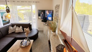 Een woonkamer met een bank, een bijzettafeltje met decoratieve objecten, een televisie en een raam met uitzicht op groen.