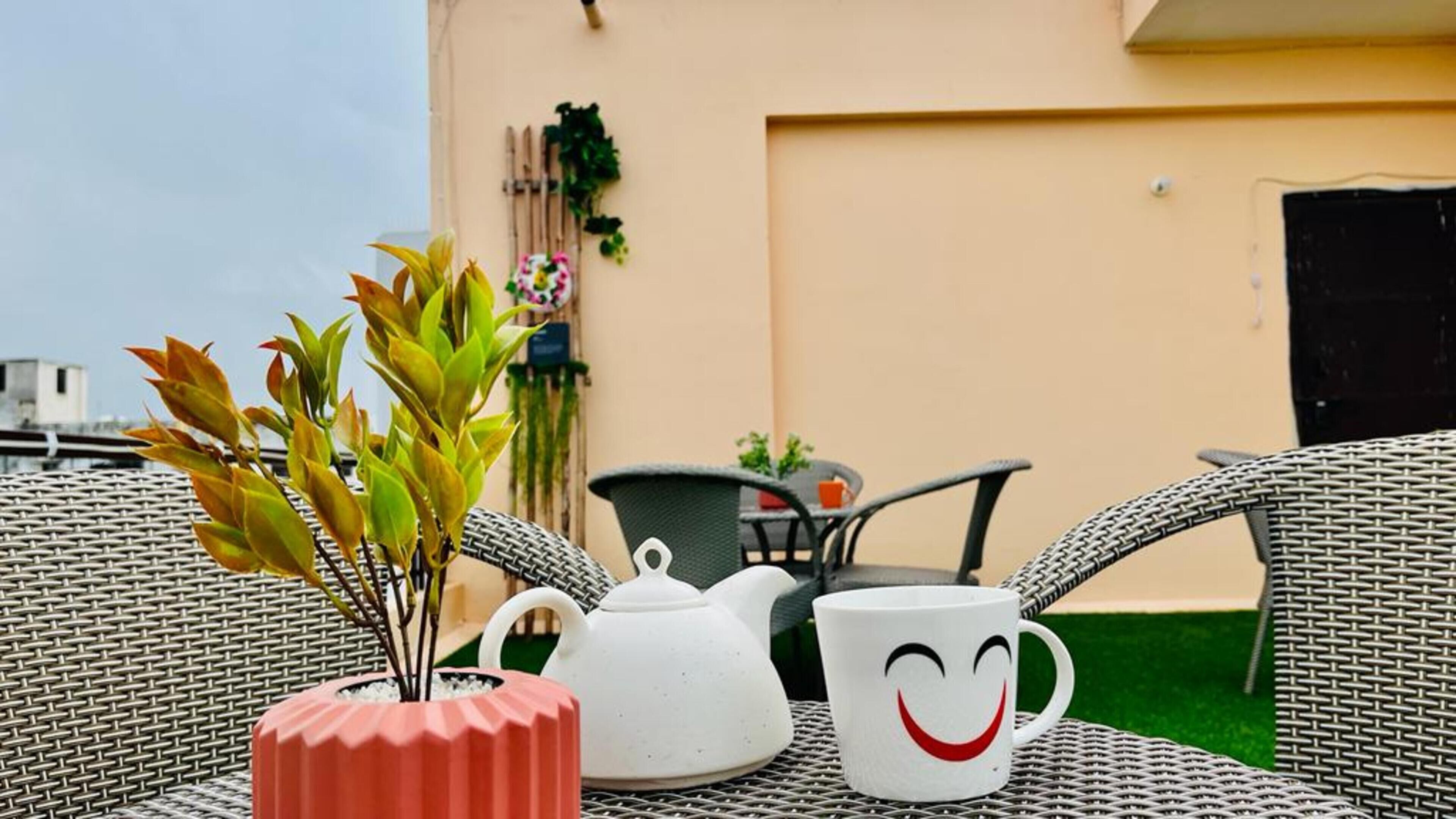 Un tavolo con teiera e tazza, circondato da sedie in vimini, in un ambiente esterno con un edificio sullo sfondo.