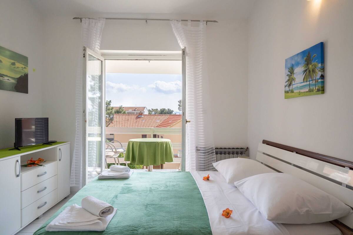 Hotellihuone, jossa on sänky, televisio, vihreä pöytä sekä parveke, jossa on pöytä ja tuolit.