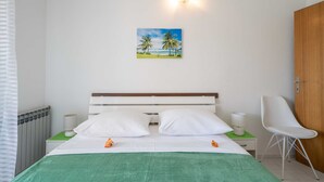 A neatly made bed with white linens, a green throw blanket, and two pillows. There is a painting of a beach scene on the wall above the bed. - Laidback Apartments (Orebic)