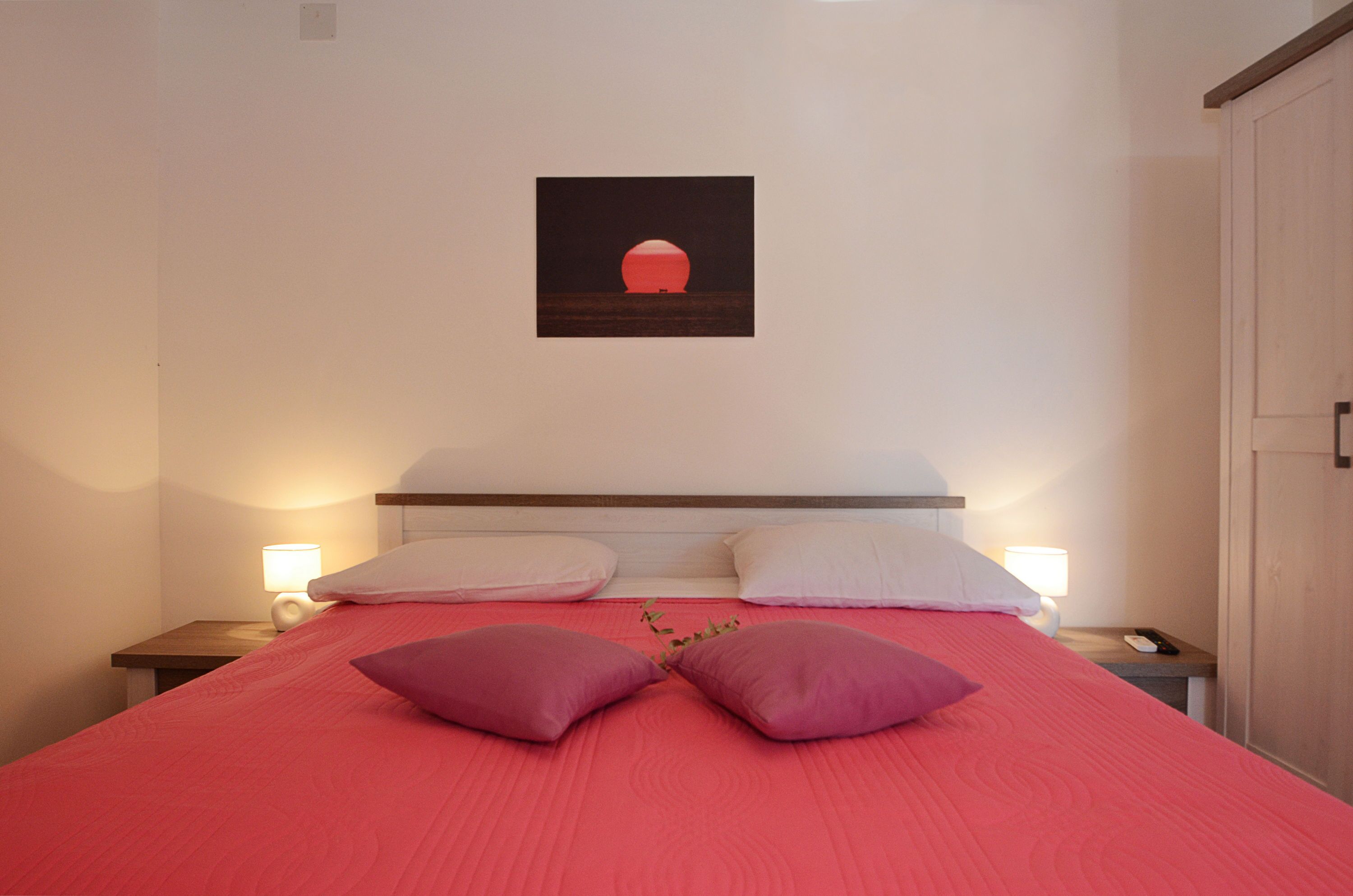 A neatly made bed with a red bedspread and two pillows, a nightstand with a lamp, and a framed picture on the wall.
