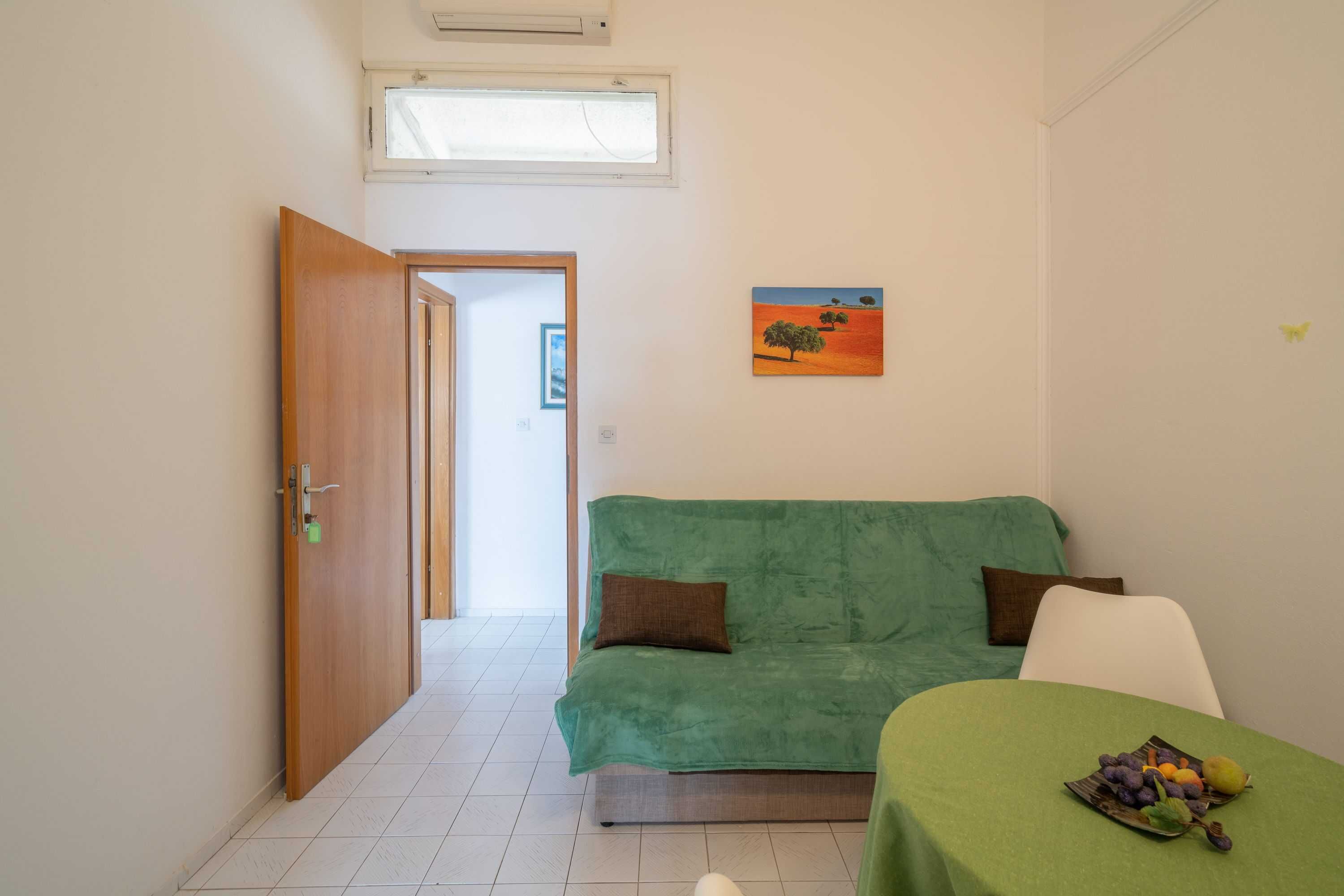 Una habitación con un sofá verde, una mesa con fruta, un cuadro y una puerta que da a otra estancia.