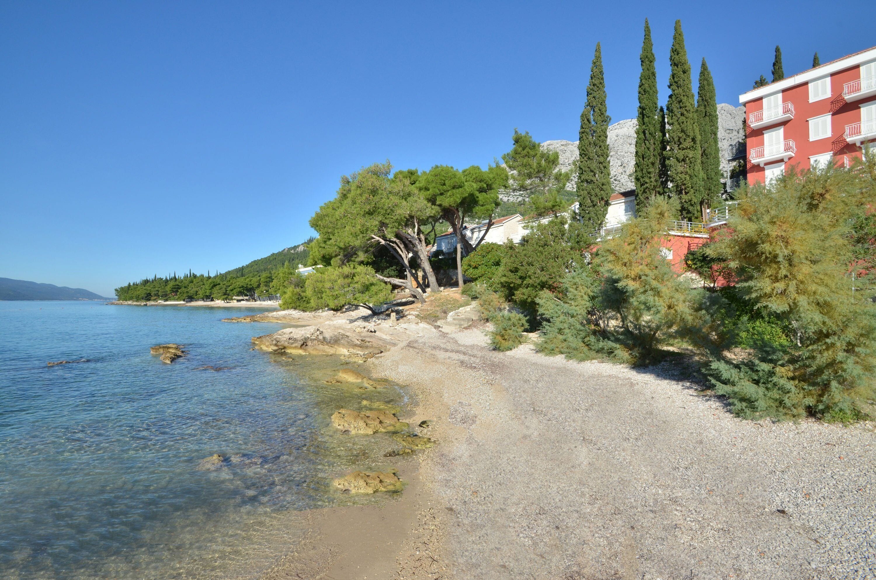 A coastal area with a clear beach, rocky shoreline, and a building with balconies overlooking the sea.