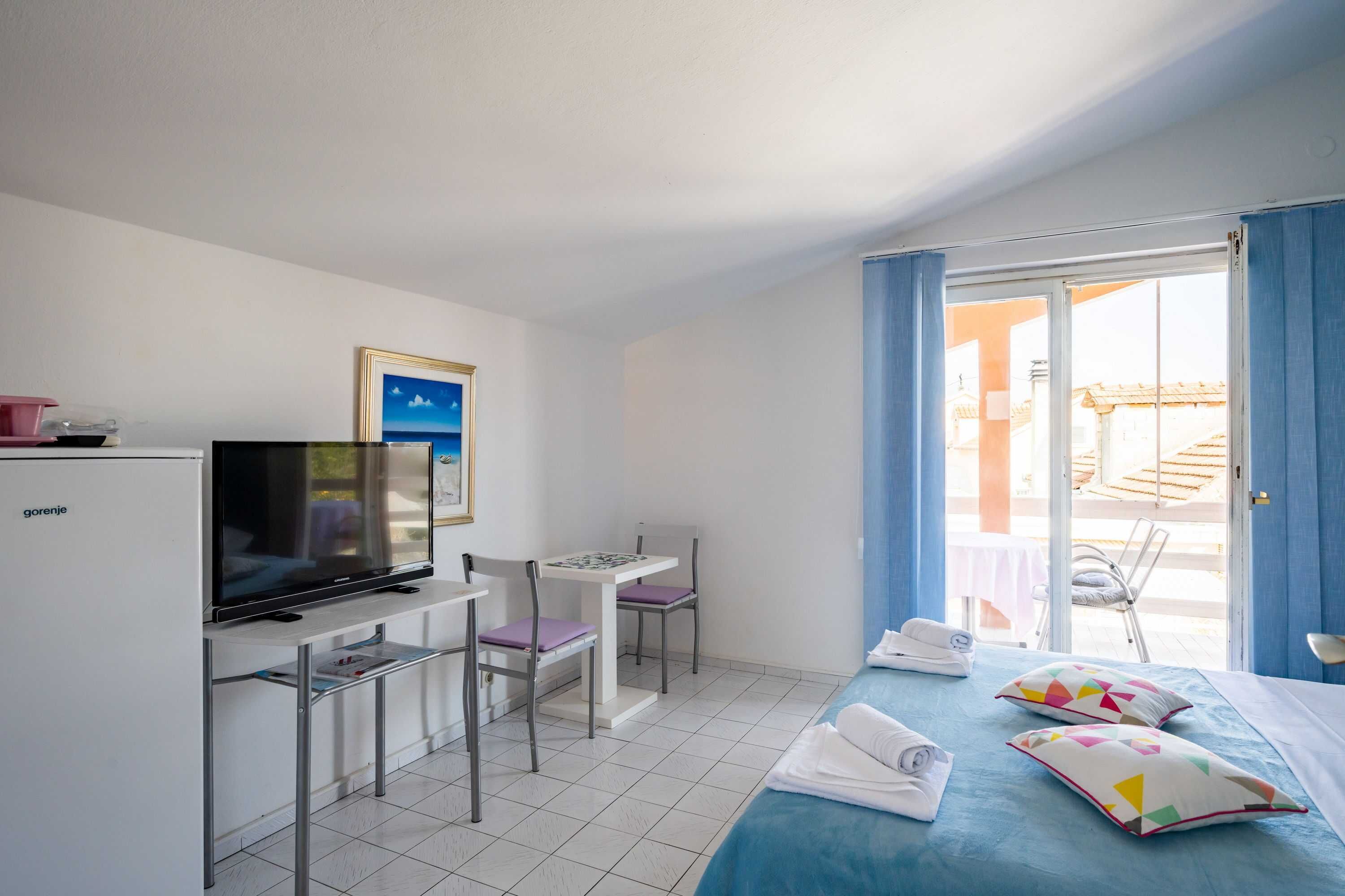 A hotel room with a bed, a desk with a television, a chair, a small table, and a window with curtains.
