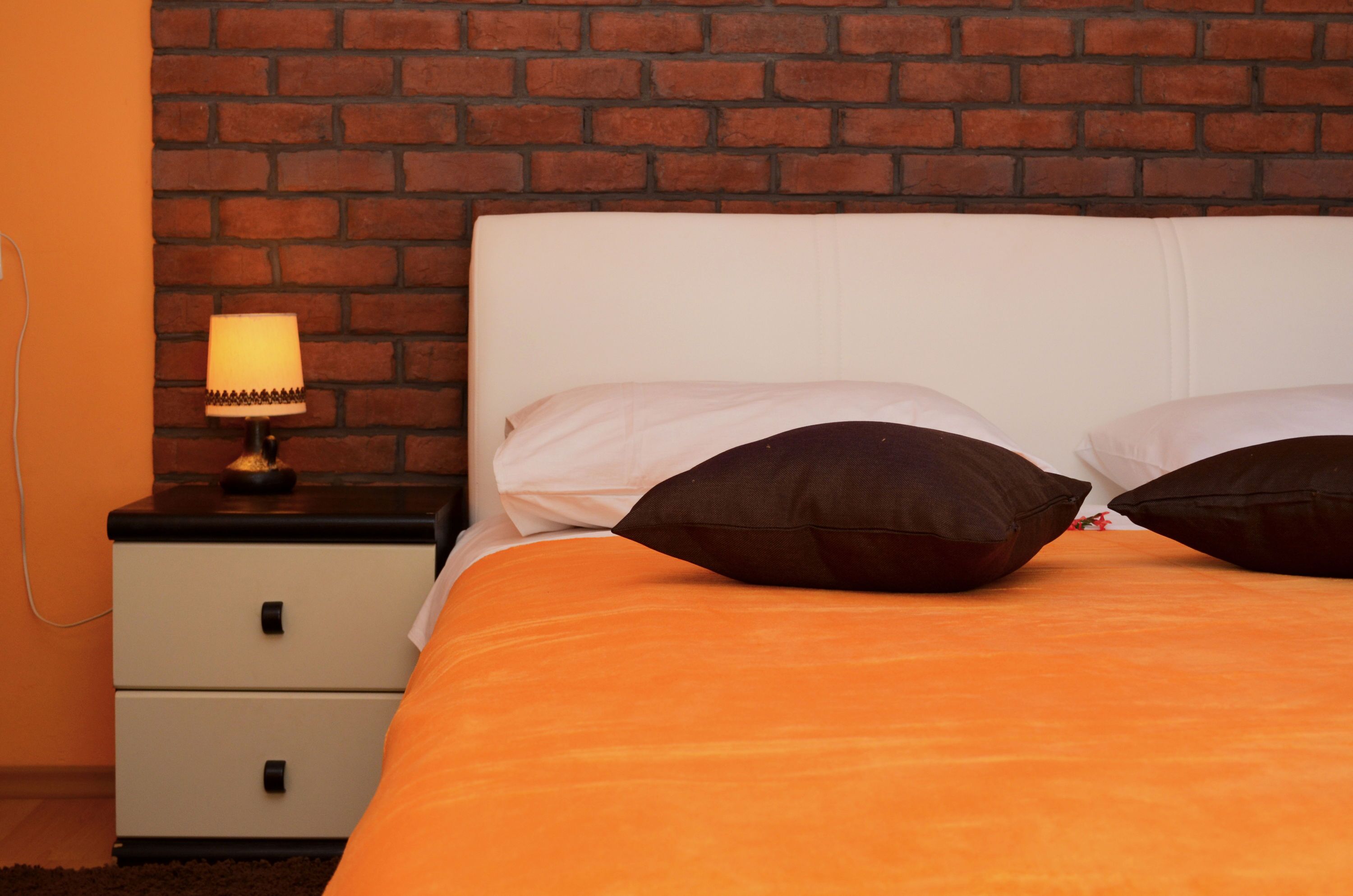 Una cama con un edredón naranja y dos almohadas oscuras, un cabecero blanco y una mesita de noche con una lámpara.
