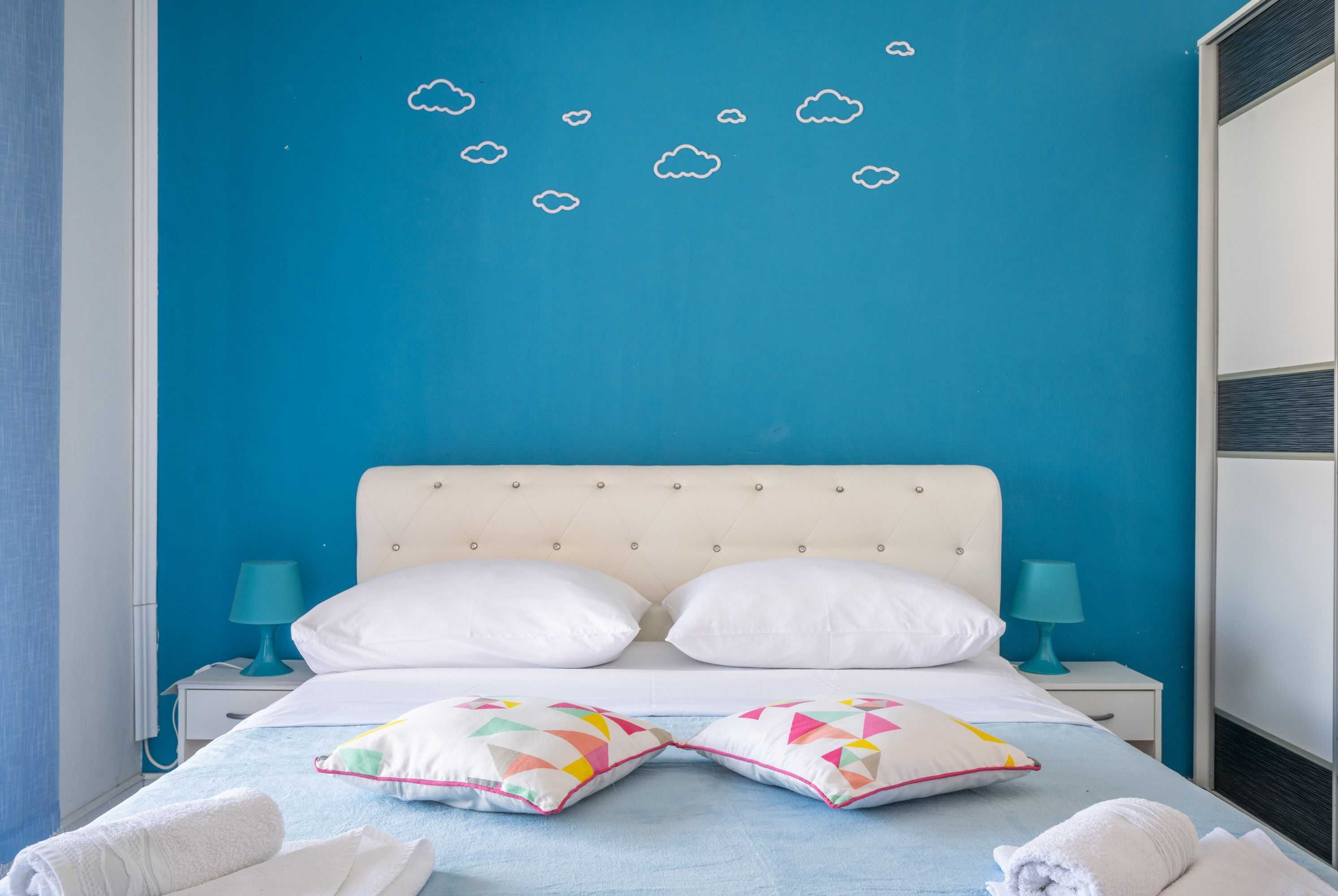 Une chambre avec un mur bleu, un lit blanc avec deux oreillers blancs, une table de chevet bleue avec une lampe, et un mur bleu décoré d’autocollants représentant des nuages.