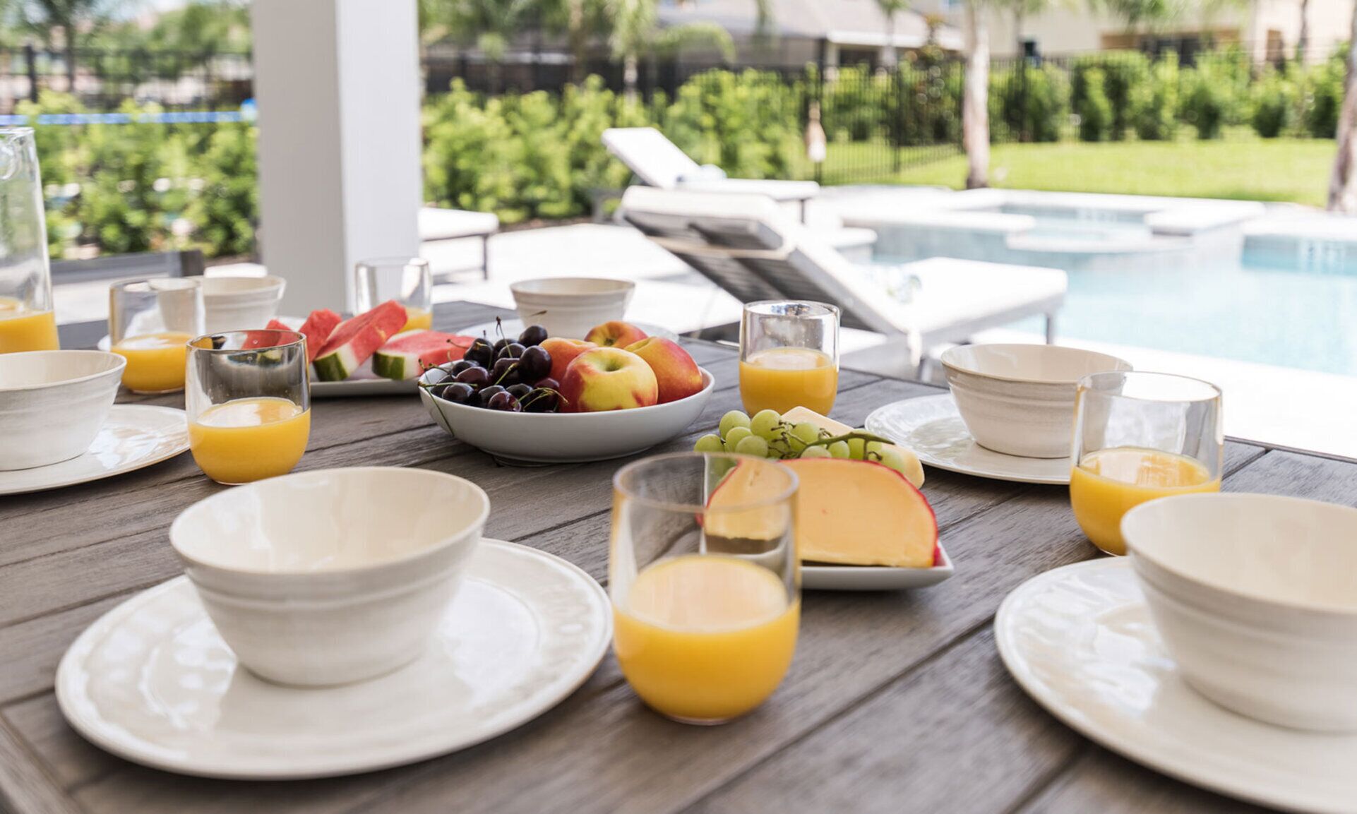 Desayuno con fruta, queso y jugo de naranja sobre una mesa de madera junto a una piscina.