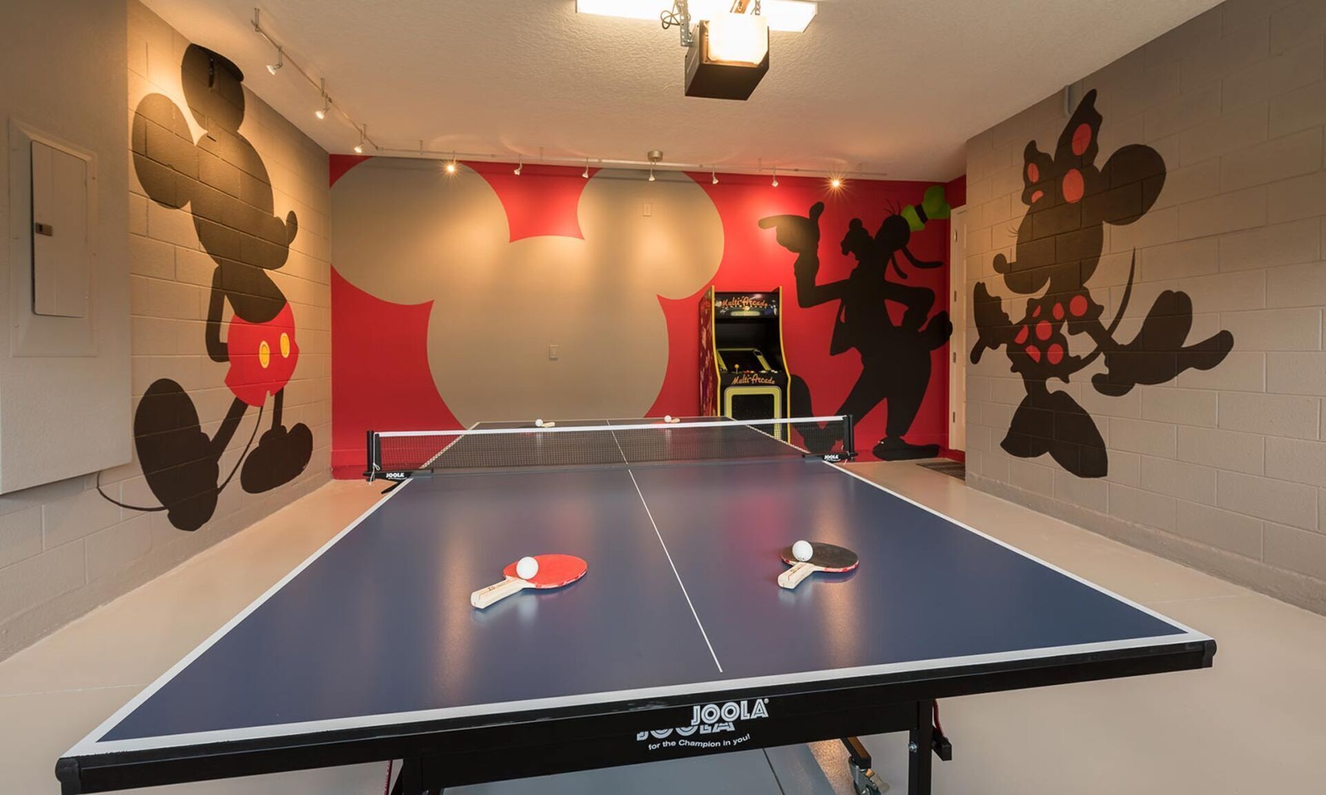 Une table de ping-pong avec deux raquettes et un filet, dans une pièce aux murs rouges et beige, avec un projecteur et une porte sur laquelle est apposée une pancarte.