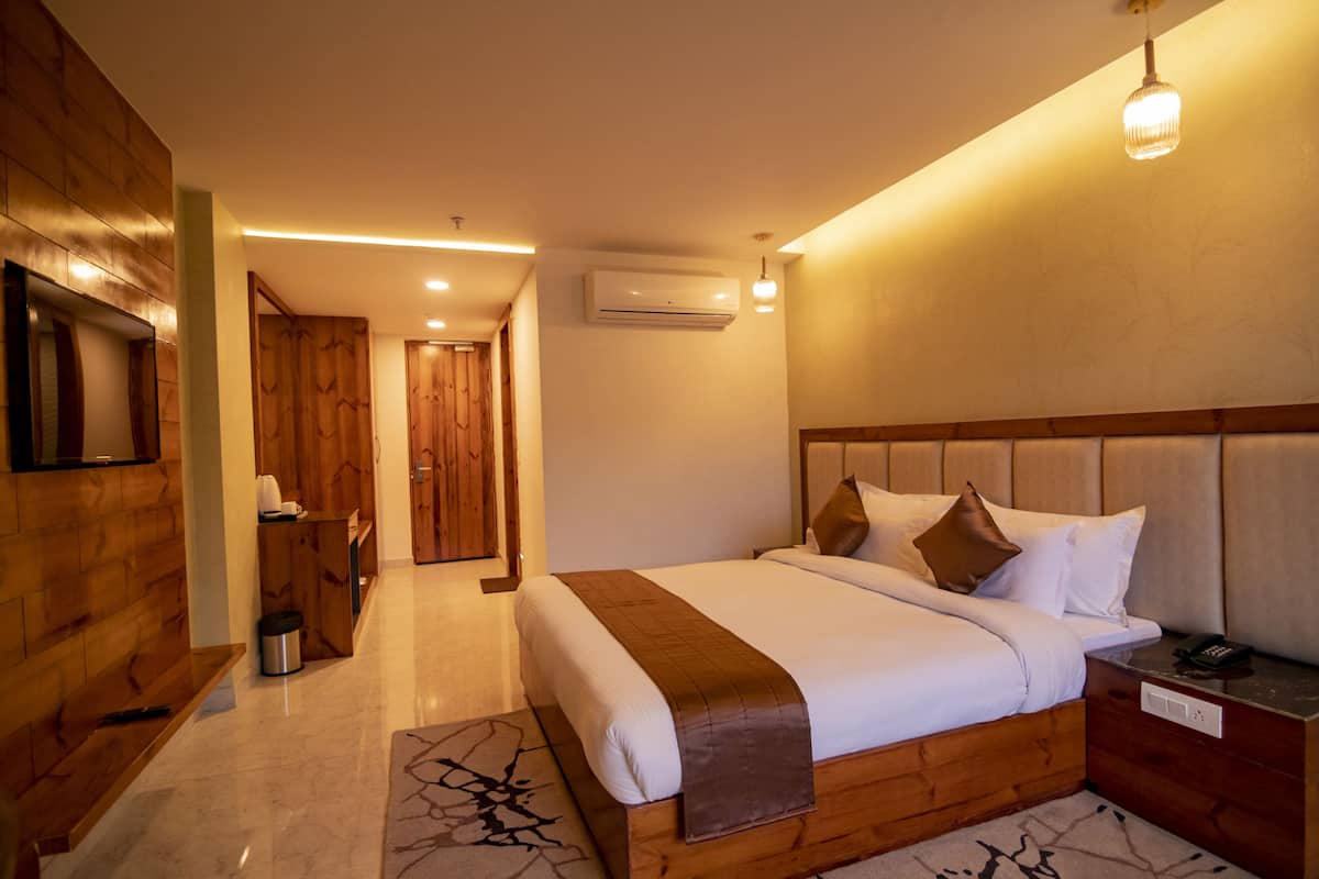 A modern hotel room with a wooden headboard, a large bed with white linens, a bedside table with a lamp, and an air conditioning unit on the wall.