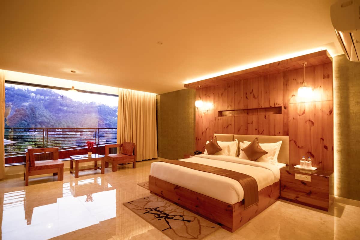 A modern bedroom with a large bed, wooden panel walls, a view of a forested area, and a balcony with chairs.