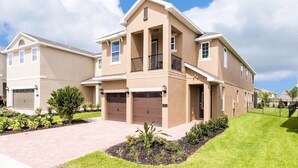 A two-story residential building with a beige exterior, a front garage, and a landscaped front yard with small shrubs and a lawn. - Rent Your Own Orlando Villa with Large Private Pool on Encore Resort at Reunion, Orlando Villa 4374 (Kissimmee)