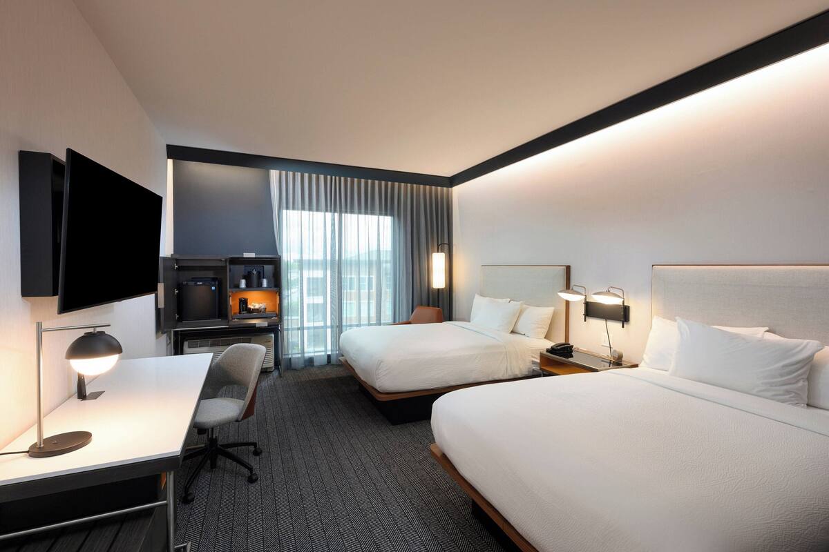Une chambre d’hôtel avec deux lits, un bureau avec un ordinateur, une télévision et un coin salon avec un fauteuil.