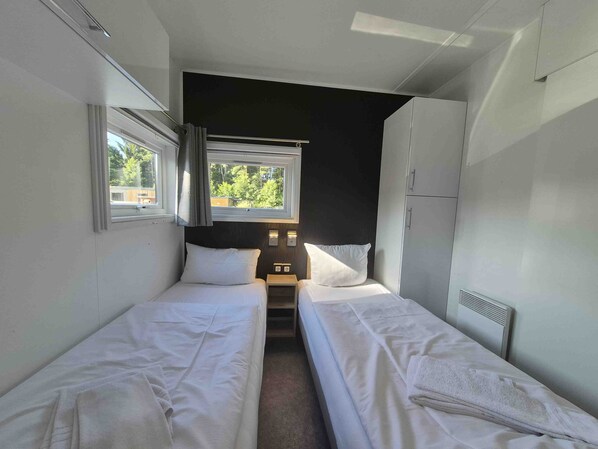 A compact room with two single beds, a small table, a window with curtains, and a radiator. - comfortable mobile home on the lake with whirlpool (WA101) (Wackersdorf)