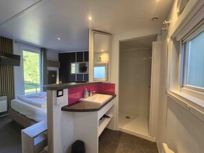 A compact hotel room with a bed, a shower, a sink, and a small window.