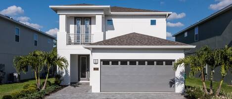 A modern two-story house with a garage, a balcony, and a well-maintained lawn with palm trees.