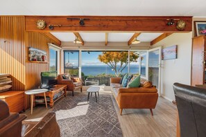 A spacious living room with a large window offering a view of the sea, wooden paneling, a comfortable sofa, and a television. - Henley - Omori Holiday Home (Omori)