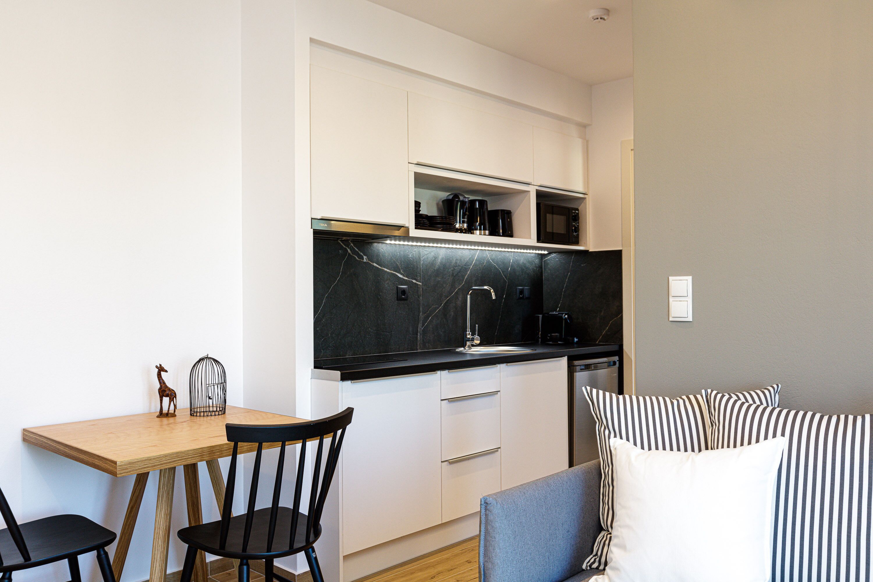 A compact kitchen with white cabinets, a black countertop, and a microwave. There is a small dining table with two chairs.