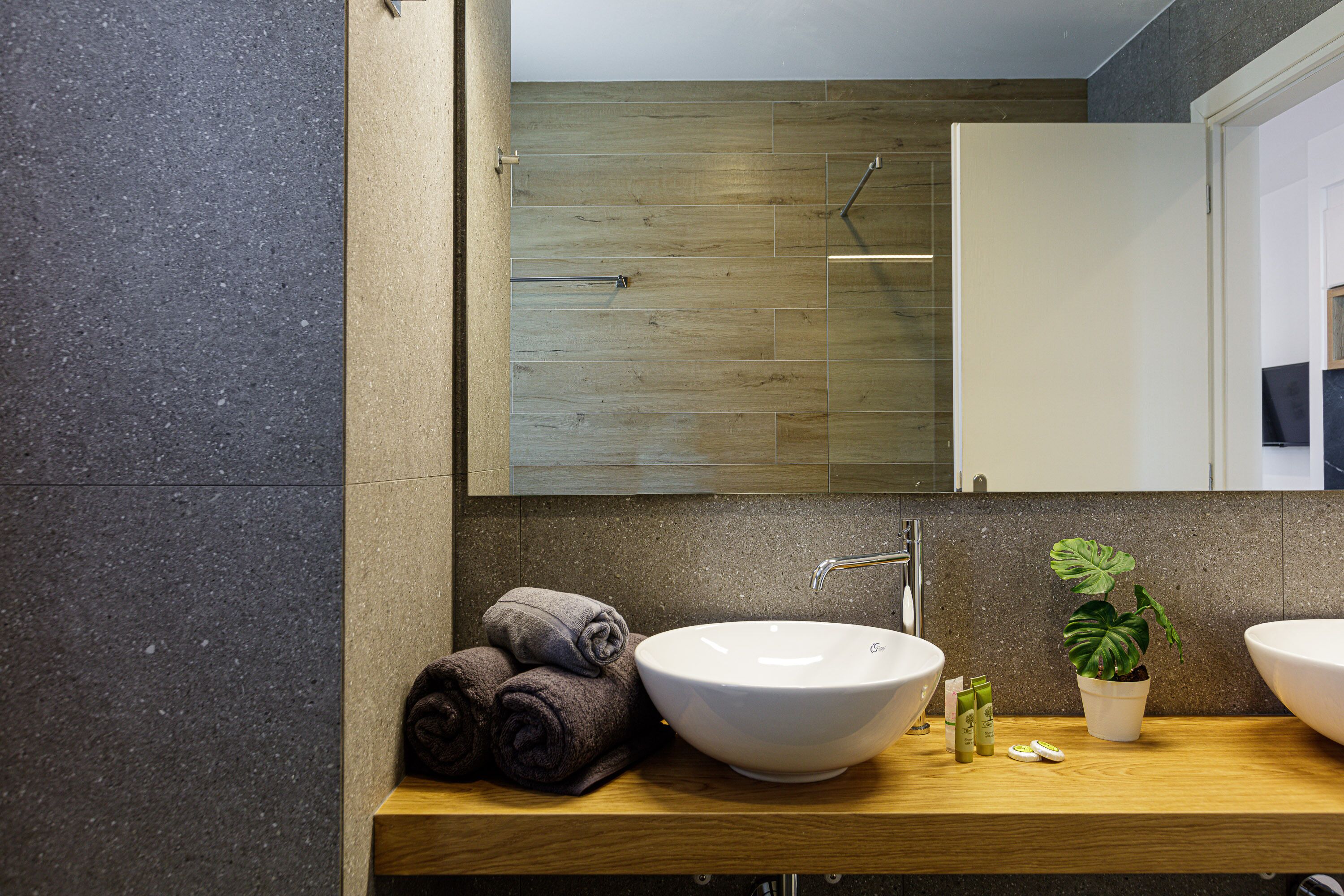 A modern bathroom with a wooden vanity, two sinks, a mirror, and a potted plant.