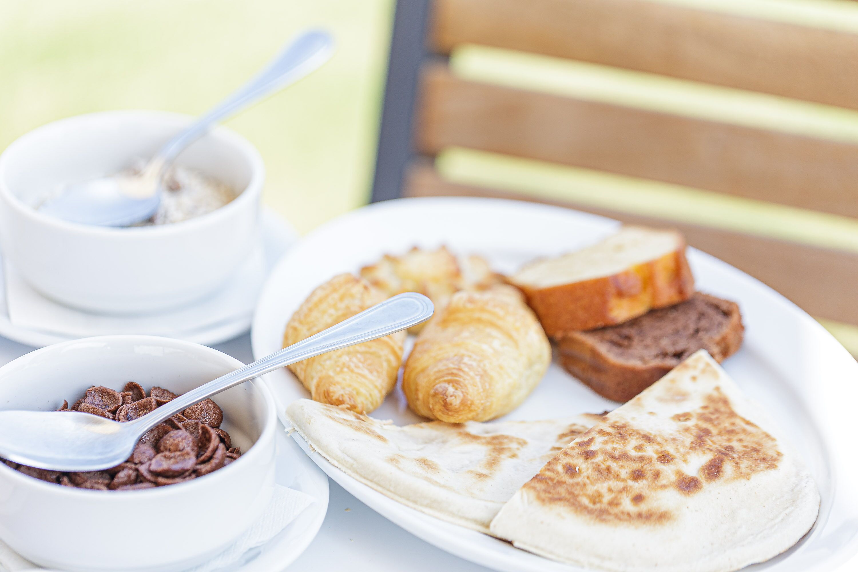 Un plato con croissants, una rebanada de pan y un tazón de cereal con una cuchara.