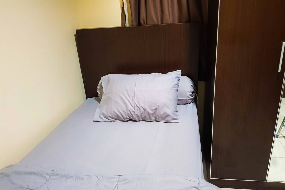 Una cama individual con cabecera, almohada blanca y edredón de tonos claros en una habitación con puerta de madera y pared lisa.