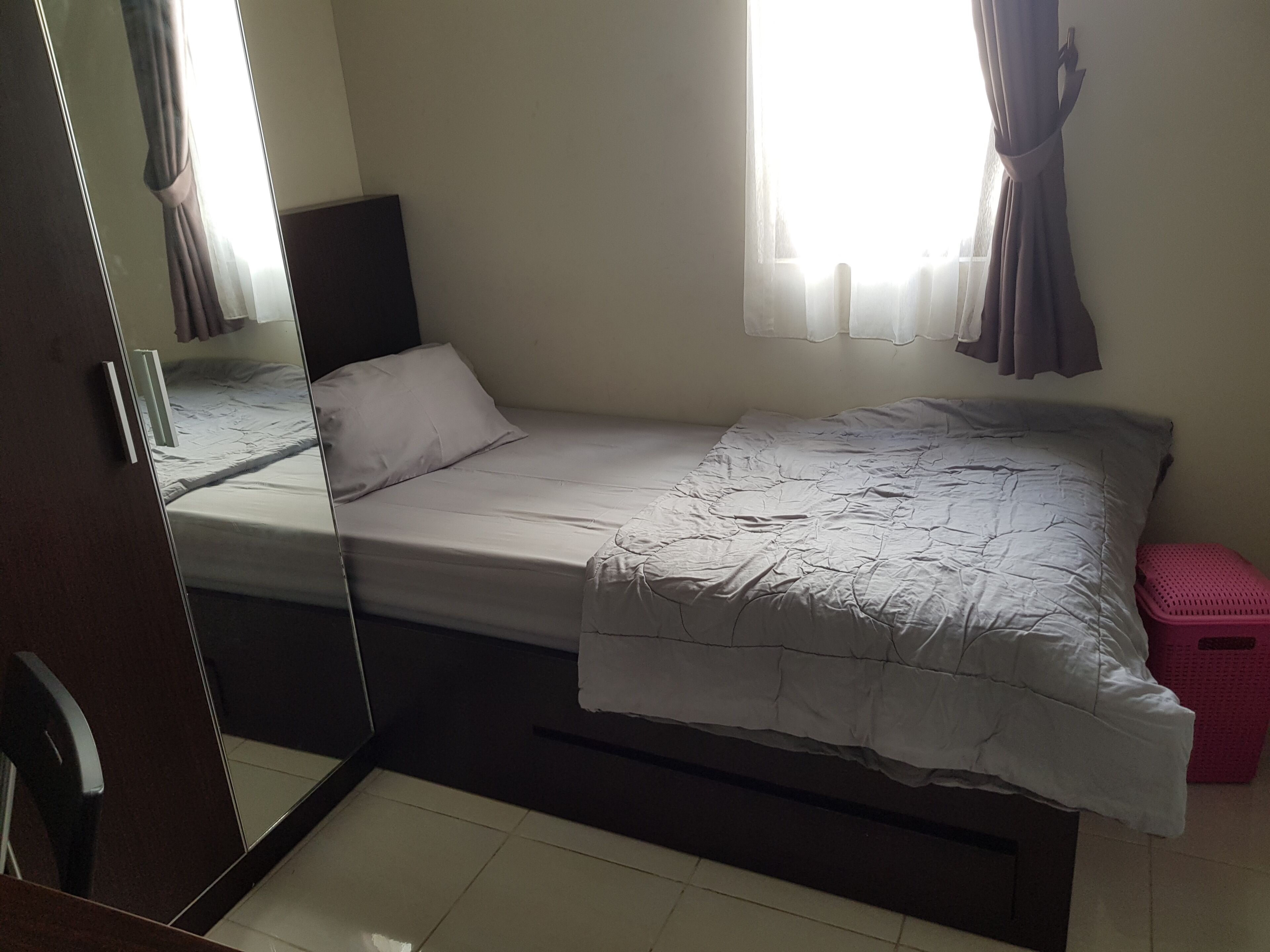 A single bed with a white mattress and a grey blanket, a small pink stool, a mirror on the wall, and a window with curtains.