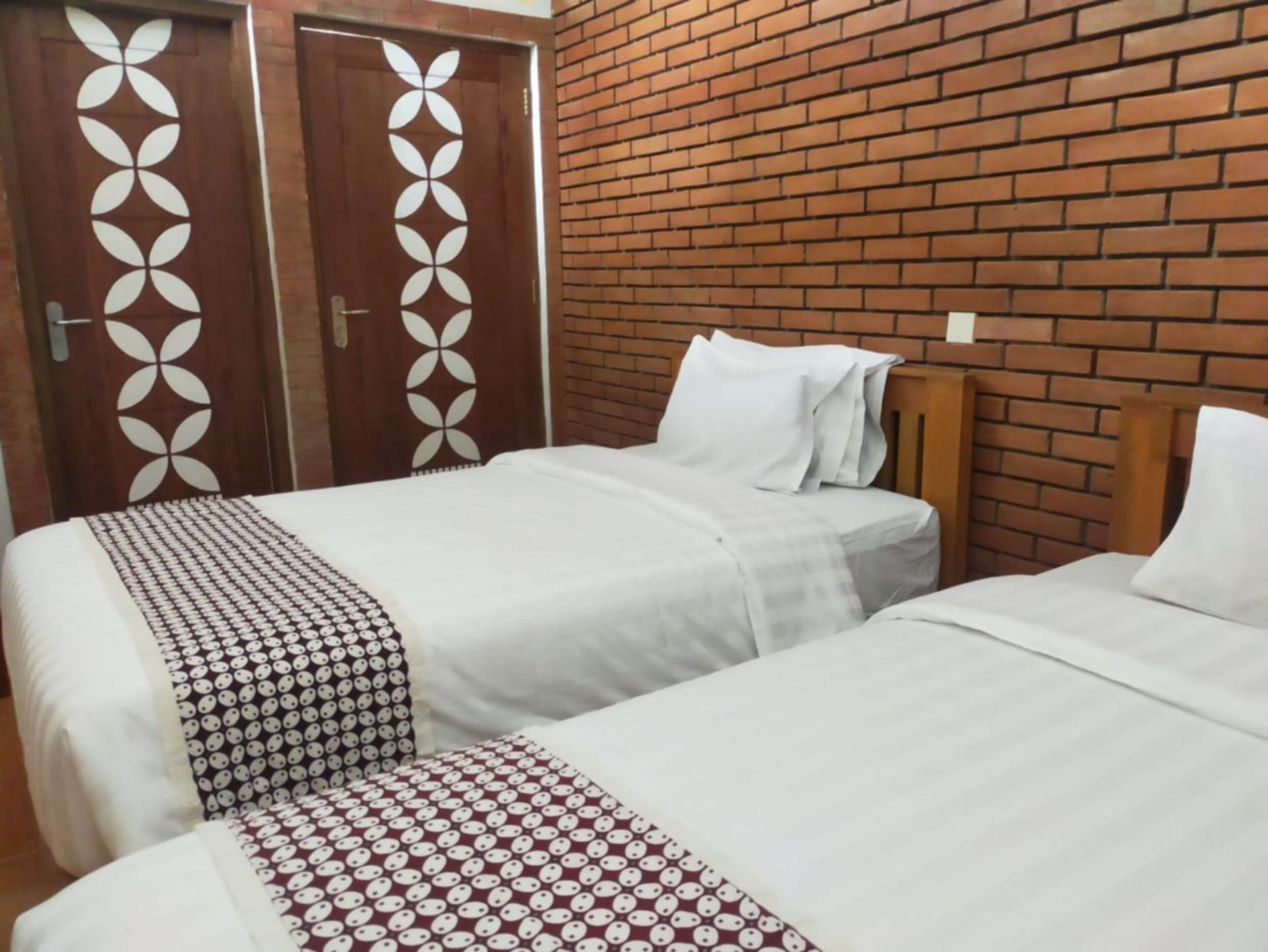A hotel room with two beds, each with white bedding and patterned throw blankets. The room features a brick wall and wooden headboard.