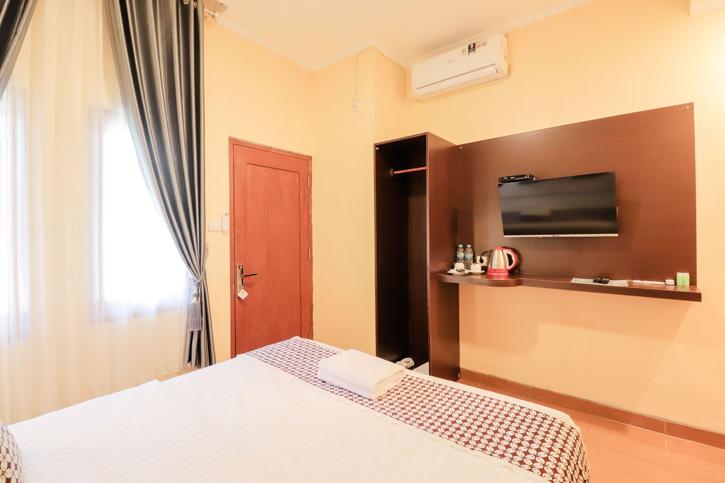 A hotel room with a bed, a television mounted on the wall, a wardrobe, and a window with curtains.