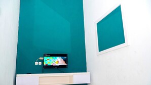 A teal-walled room with a mounted TV on a white shelf, a window, and a white door. - Kozi Kost Paviliun (Tegal)