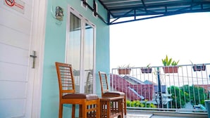 A balcony with two wooden chairs, a metal railing, and a view of a red-roofed building in the background. - Kozi Kost Paviliun (Tegal)