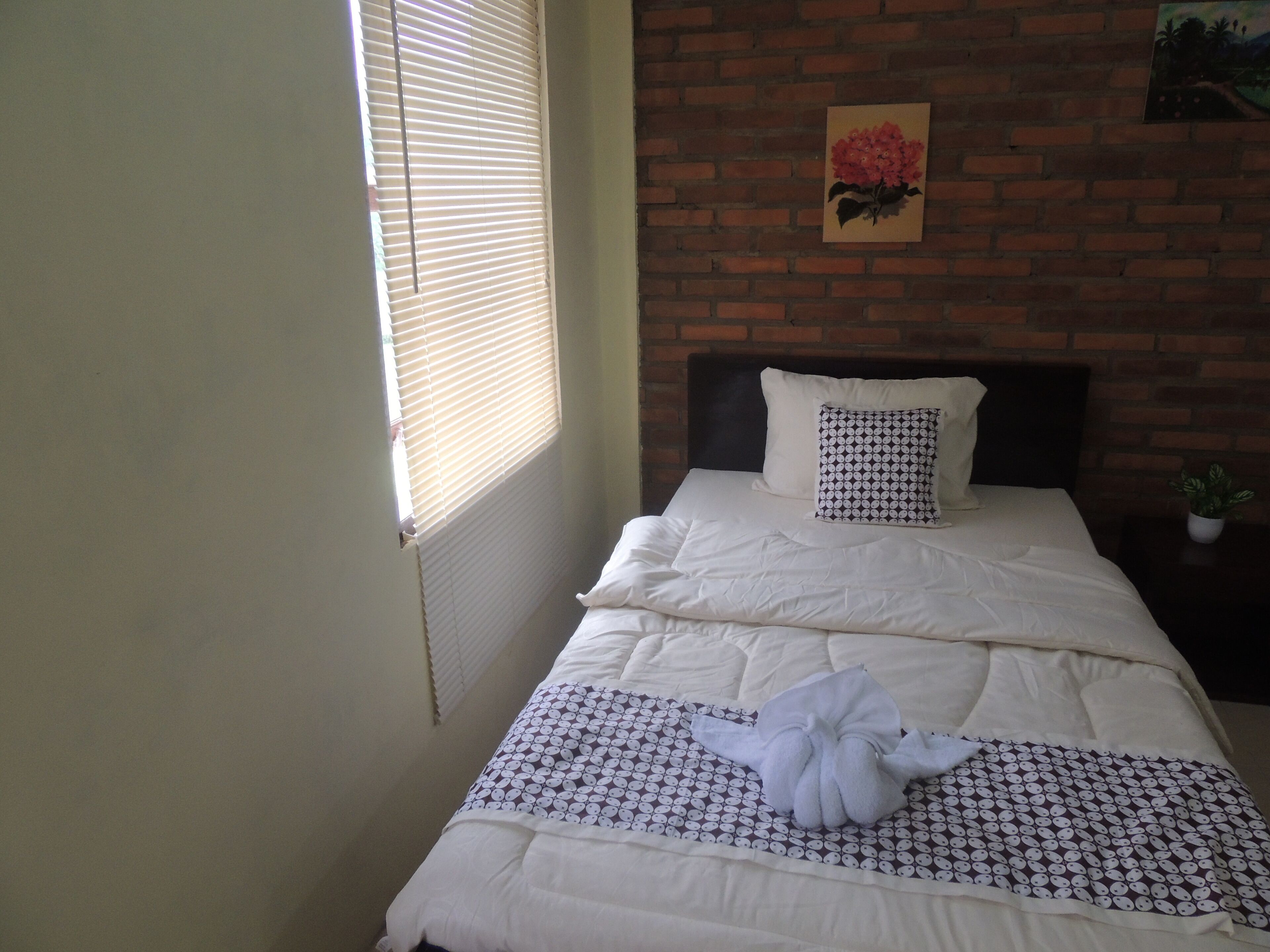 A neatly made bed with white bedding and a patterned pillow, a small potted plant on a nightstand, and a brick wall with a framed picture.