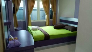 A single-bed room with a green bedspread, a brown headboard, and a window with curtains.