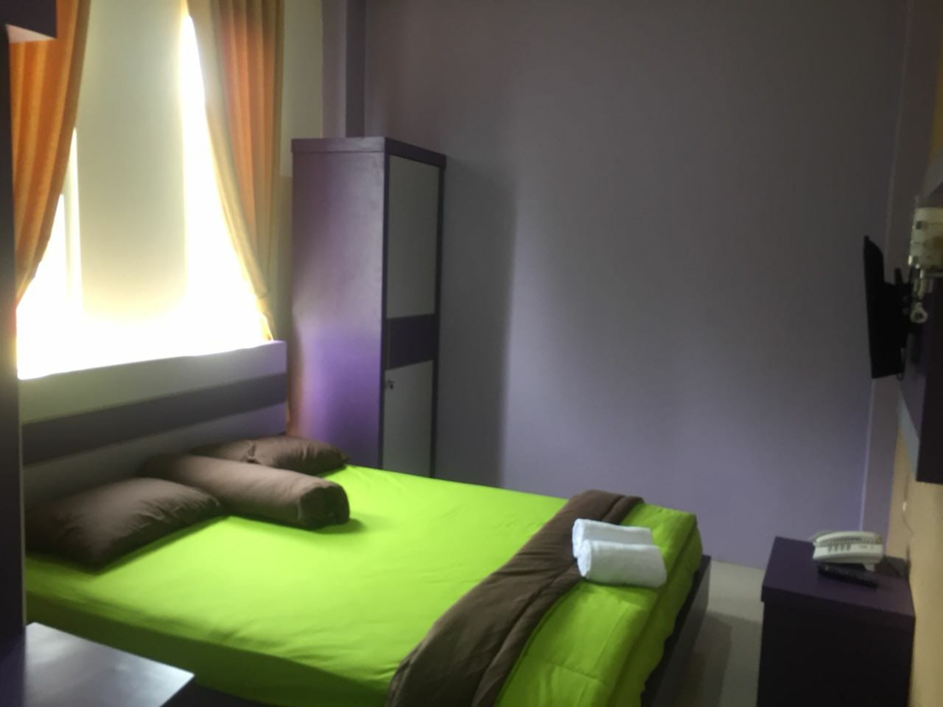 A single bed with green bedding, a brown pillow, and a white towel. There is a purple cabinet against the wall and a small table with a remote control on it.