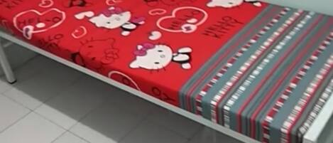 Una cama individual con una colcha temática de Hello Kitty en rojo en una habitación con suelo de baldosas blancas.