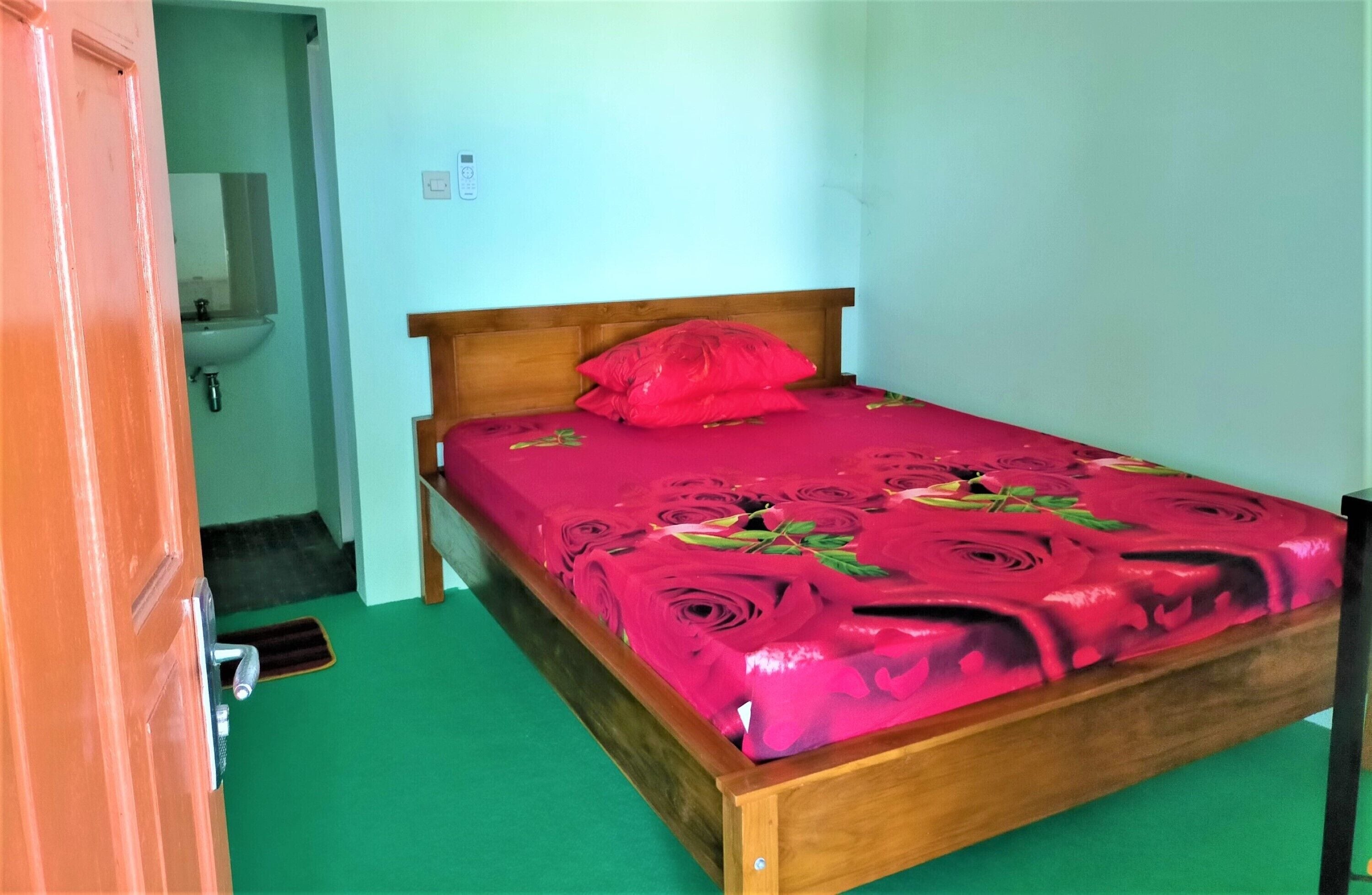 Una habitación pequeña con cama individual, cabecera de madera, colcha estampada de flores rojas y alfombra verde.