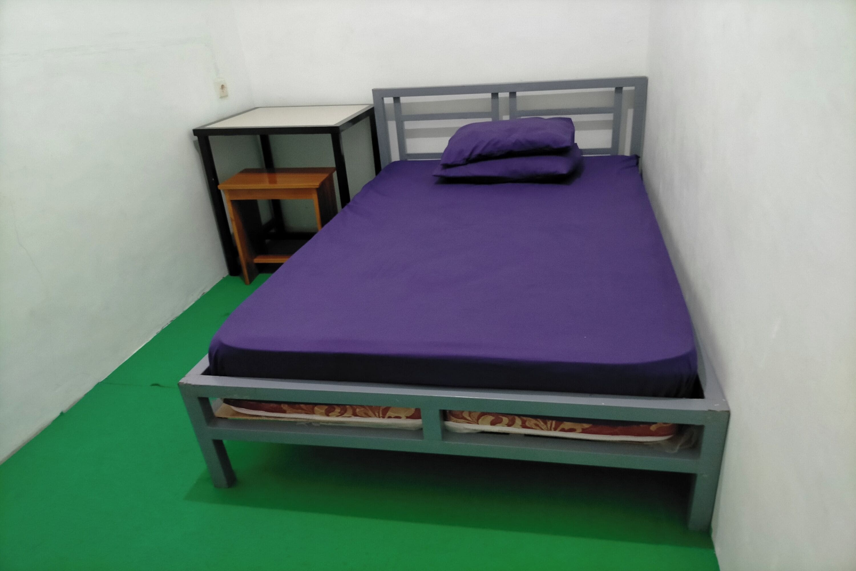 A single bed with a purple mattress and pillows, a wooden nightstand, and a green floor.