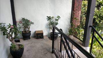 Een betonnen trap met zwarte leuningen leidt naar een balkon. Op het balkon staan potplanten, waaronder een grote met roze bloemen en enkele kleinere.