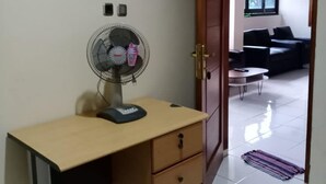 Ein Zimmer mit Schreibtisch, Standventilator, einer zur anderen Kammer führenden Tür und einem Fenster.