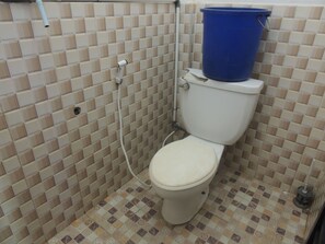 Une petite salle de bain avec des toilettes, un seau bleu posé sur le réservoir des toilettes, et des murs et un sol carrelés.