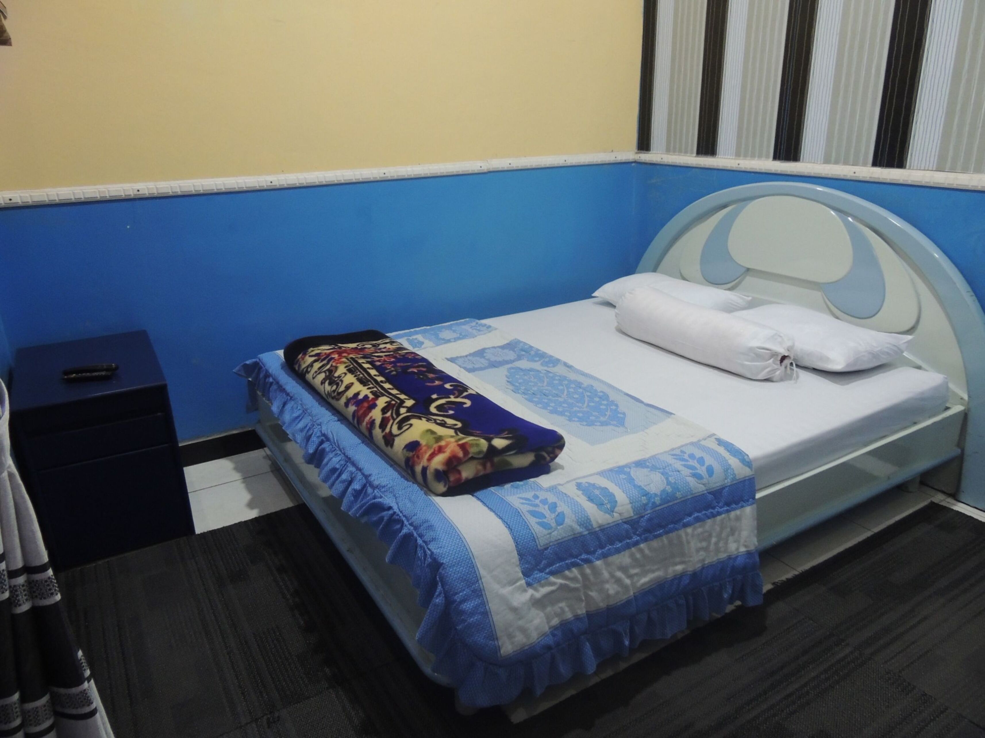 A single bed with a blue and white bedspread, a decorative pillow, and a small bedside table with a remote control.